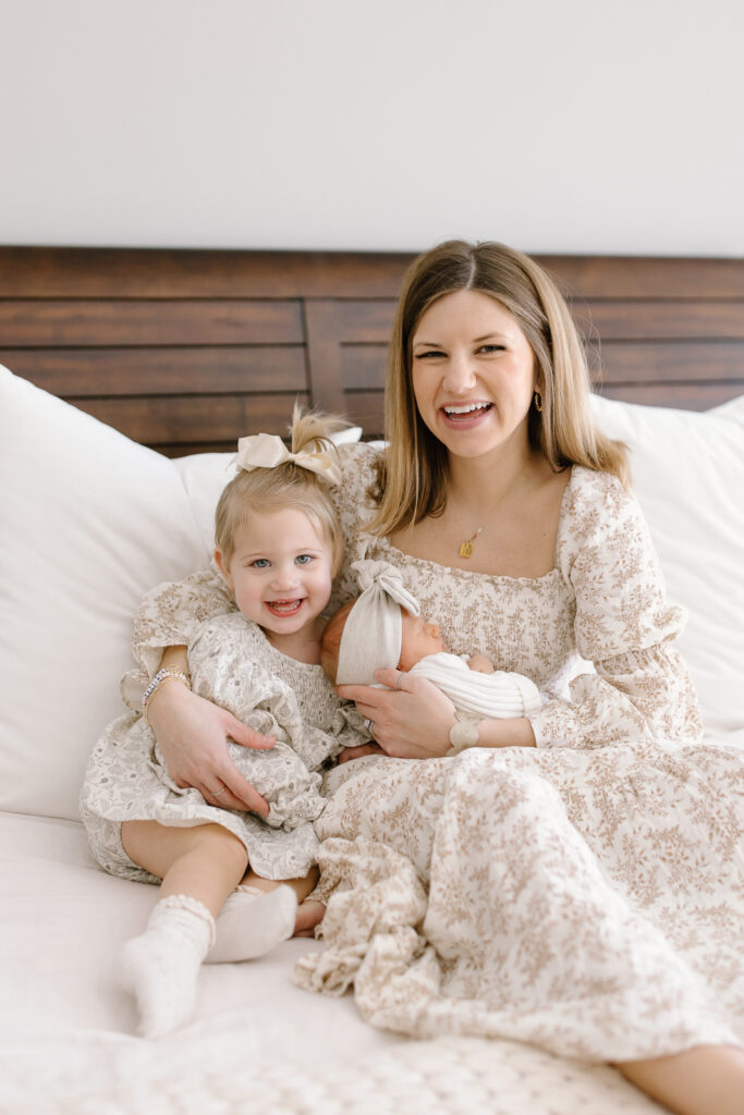 Mom cuddling in bed with her two kids and smiling towards the camera during indianapolis newborn photos