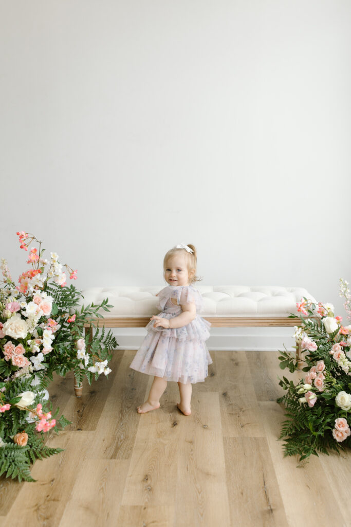 Little girl standing at bench and smiling towards the camera during Mother's Day Mini Sessions at Aspiring Eden Studio in Indianapolis