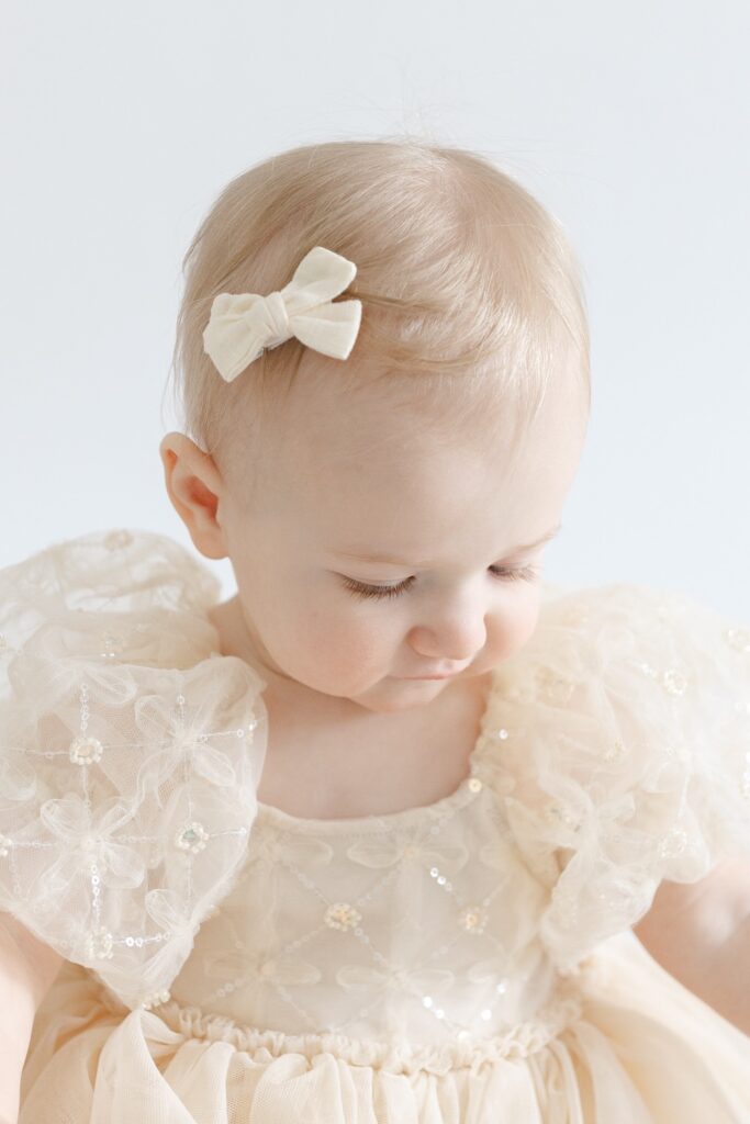 Little girl looking down while wearing a tan dress and tan bow during first birthday photos in Indianapolis with Capturing Simplicity Photography