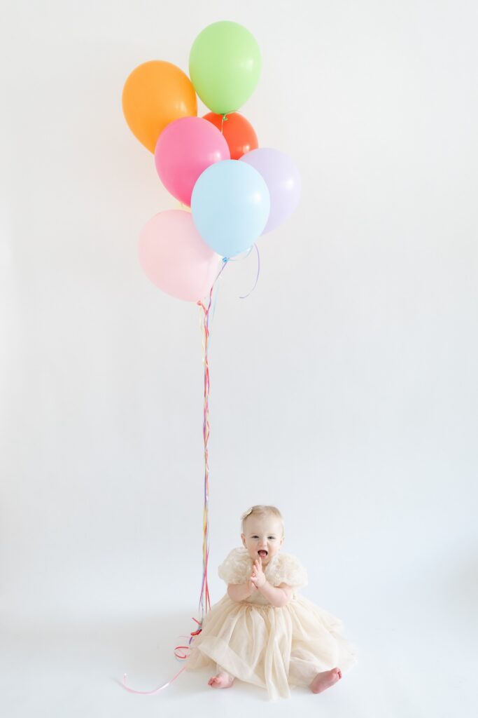 Baby girl sitting and clapping with colorful balloons int he background during first birthday photos in Indianapolis with Capturing Simplicity Photography